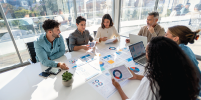 Multi racial group of people working with Paperwork on a board room table at a business presentation or seminar. The documents have financial or marketing figures, graphs and charts on them. There is a laptop and digital tablet on the table. Multi ethnic group including Caucasian and African American