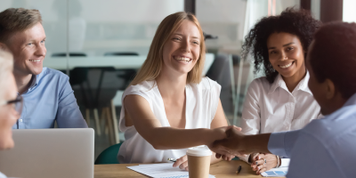 A young business woman shaking hands with a co-worker surrounded by others - Interview Skills for Applicants - Behavioral Based Interviewing for Interviewers