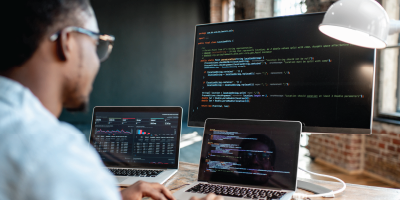 Young african male programmer writing program code sitting at the workplace with three monitors in the office. Image focused on the screen