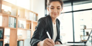 A professional woman taking notes on a notepad while working at her computer.