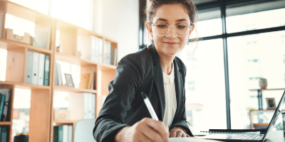 A professional woman taking notes on a notepad while working at her computer.